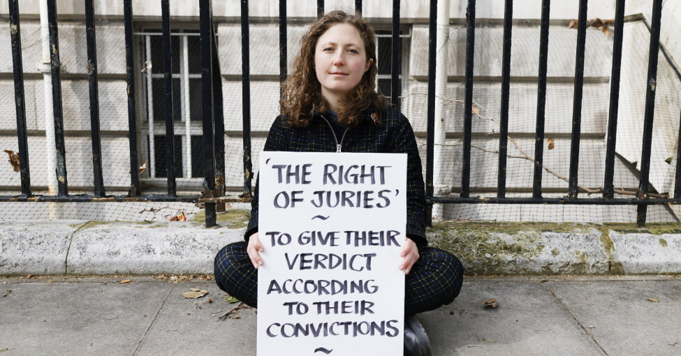 Indigo Rumbelow sitting cross legged and holding a sign reading 'The right of juries ~ to give their verdict according to their convictions'